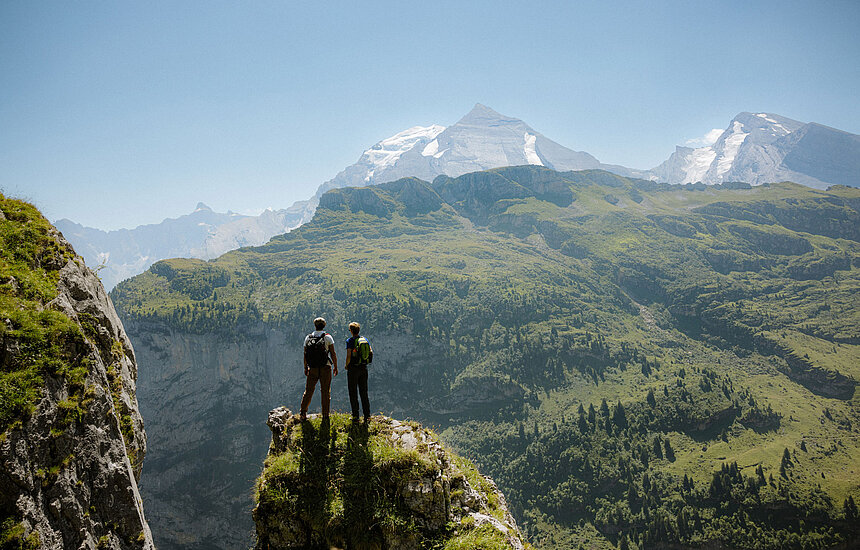 <p>Zwei Wanderer stehen auf einem Felsvorsprung und betrachten die grüne Berglandschaft mit imposanten Gipfeln.</p>